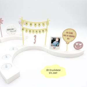 May include: A white, curved display with birthday decorations. Includes a banner that reads "HAPPY BIRTHDAY", a photo, a balloon, and a smiley face. The number "3" is also displayed. The display is decorated with pink and yellow accents.