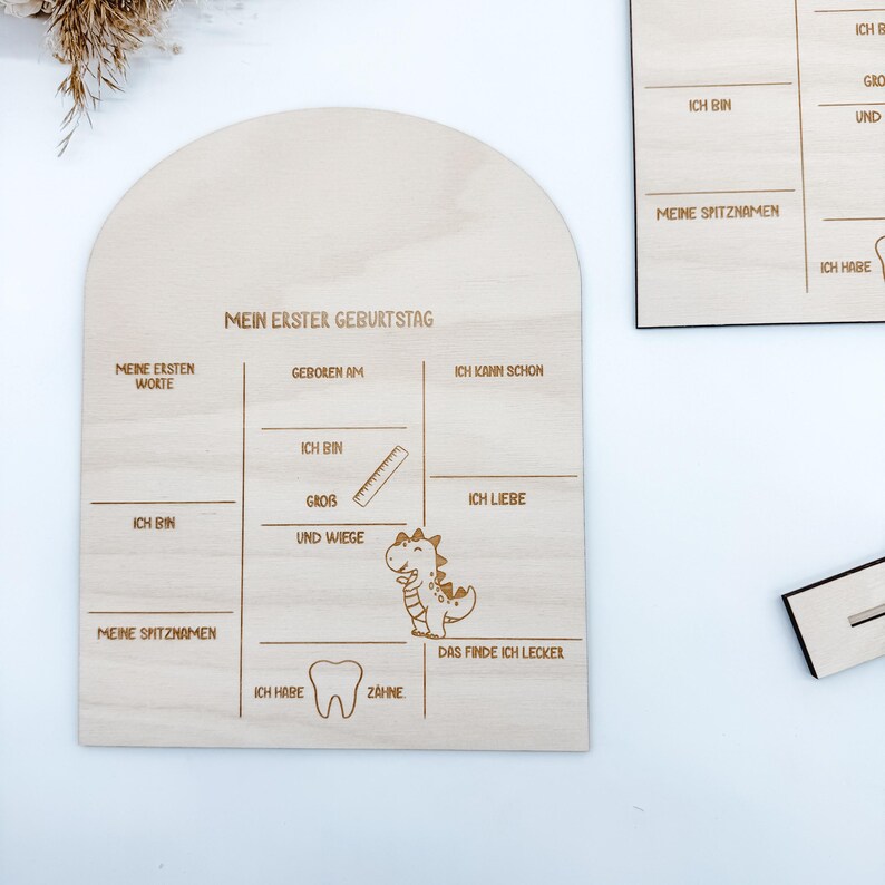 Puede incluir: Un letrero de madera con un gr&aacute;fico de dinosaurio y el texto "Mi primer cumplea&ntilde;os". El letrero tiene espacios para completar informaci&oacute;n sobre el primer a&ntilde;o del ni&ntilde;o, como sus primeras palabras, cu&aacute;nto pesa y qu&eacute; le gusta.