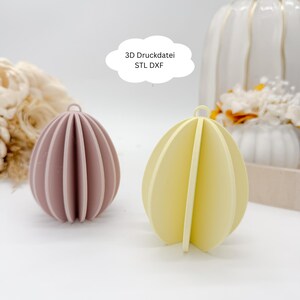 Puede incluir: Dos adornos decorativos en forma de huevo. Uno es de color malva apagado y el otro es amarillo suave. Ambos tienen un diseño segmentado con un pequeño lazo en la parte superior. Un gráfico blanco en forma de nube sobre los adornos dice "3D Druckdatei STL DXF".
