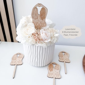 Op de afbeelding: Twee lasergesneden houten bloemstekers met een ontwerp van twee meisjes die elkaars hand vasthouden. De stekers staan in een witte keramische vaas met witte en roze bloemen.