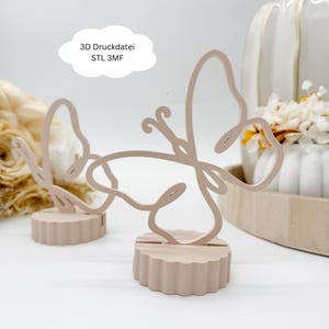 Puede incluir: Dos decoraciones en forma de mariposa rosa claro. Las mariposas están hechas de una línea fina y continua y se asientan sobre una base festoneada. El texto "3D Druckdatei STL 3MF" está en un cuadro de texto blanco en forma de nube.