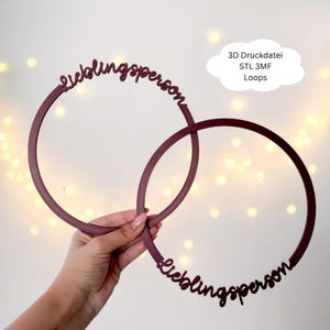 May include: Two burgundy-colored circular wall hangings with the word "Lieblingsperson" in script. The text "3D Druckdatei STL 3MF Loops" is in a white cloud-shaped text box. The background has soft, out-of-focus lights.