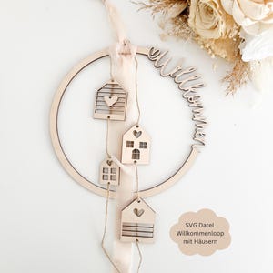 以下が含まれることがあります： 筆記体で「Willkommen」と書かれた木製の円形リース。4つの小さな木製の家の切り抜きが、ひもで円から吊り下げられています。家にはハートと窓のディテールがあります。上部には薄いピンクのリボンが付いています。