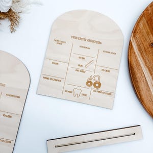 Puede incluir: Un letrero de madera con el texto "Mein erster Geburtstag" (Mi primer cumplea&ntilde;os) y espacios para rellenar informaci&oacute;n sobre el primer cumplea&ntilde;os de un ni&ntilde;o, incluyendo sus primeras palabras, su peso y lo que le gusta comer. El letrero est&aacute; decorado con un tractor y un diente.