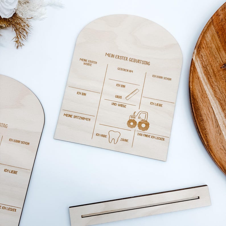 Puede incluir: Un letrero de madera con el texto "Mein erster Geburtstag" (Mi primer cumplea&ntilde;os) y espacios para rellenar informaci&oacute;n sobre el primer cumplea&ntilde;os de un ni&ntilde;o, incluyendo sus primeras palabras, su peso y lo que le gusta comer. El letrero est&aacute; decorado con un tractor y un diente.