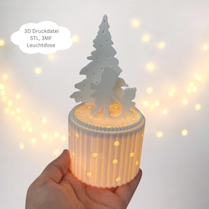 May include: A white LED lamp with a cylindrical base and a decorative top featuring a Christmas tree and two figures. The lamp has vertical grooves and small, round cutouts that emit a warm glow. Text on the image reads: "3D Druckdatei STL, 3MF Leuchtdose."