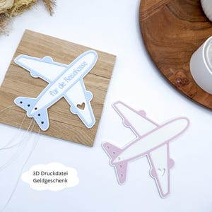 May include: Two airplane-shaped decorations, one light blue and one pink, with white accents. The light blue plane has the text "für die Reisekasse" and is mounted on a wooden square. The pink plane is on a white surface. The text "3D Druckdatei Geldgeschenk" is also visible.