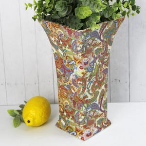 May include: A decorative vase with a colorful paisley pattern in red, blue, and green. The vase is filled with green leafy branches and has a gold rim. A yellow lemon rests beside the vase.