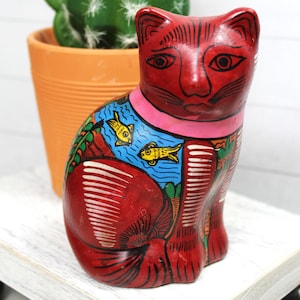 May include: A red ceramic cat figurine with a pink collar and painted details. The cat has a blue section with yellow fish and green and white accents. A green cactus and orange pot are in the background.