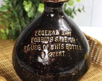 May include: A dark brown ceramic bottle with a wide base and narrow neck, featuring embossed text that reads "FEDERAL LAW FORBIDS SALE OR RE-USE OF THIS BOTTLE". The bottle contains green plant stems. A decorative piece.