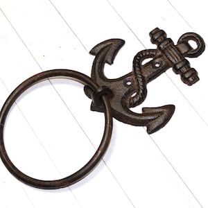 May include: A rustic, cast iron anchor-shaped towel ring. The anchor is dark brown with a rope detail and a circular ring for hanging towels. The nautical-themed design is set against a white wooden background.