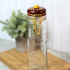 Puede incluir: Un tarro de vidrio transparente con una tapa marrón y amarilla. El tarro tiene un cierre de alambre y una superficie texturizada. El tarro está sentado sobre una tabla de cortar de madera.