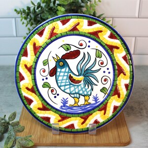 May include: A white ceramic plate with a colorful rooster design. The rooster is blue, yellow, and red with a green and yellow border around the plate. The plate has a mosaic-like design.
