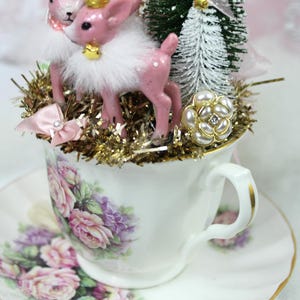 May include: A decorative teacup and saucer set featuring a floral design. The teacup is filled with a festive arrangement of pink deer figurines, miniature Christmas trees, and gold tinsel. A pearl and rhinestone embellishment adds a touch of elegance.