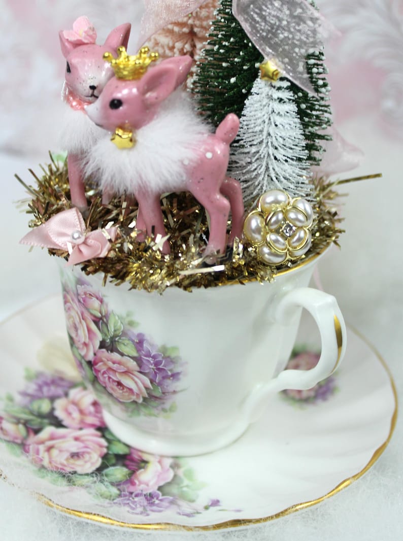 May include: A decorative teacup and saucer set featuring a floral design. The teacup is filled with a festive arrangement of pink deer figurines, miniature Christmas trees, and gold tinsel. A pearl and rhinestone embellishment adds a touch of elegance.