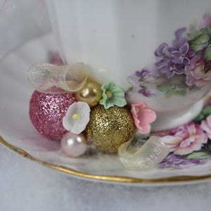 May include: A white teacup and saucer set with a floral design in pink and purple. The cup is decorated with glittery pink and gold embellishments, small white and pink flowers, and a pearl. The saucer has a gold rim.