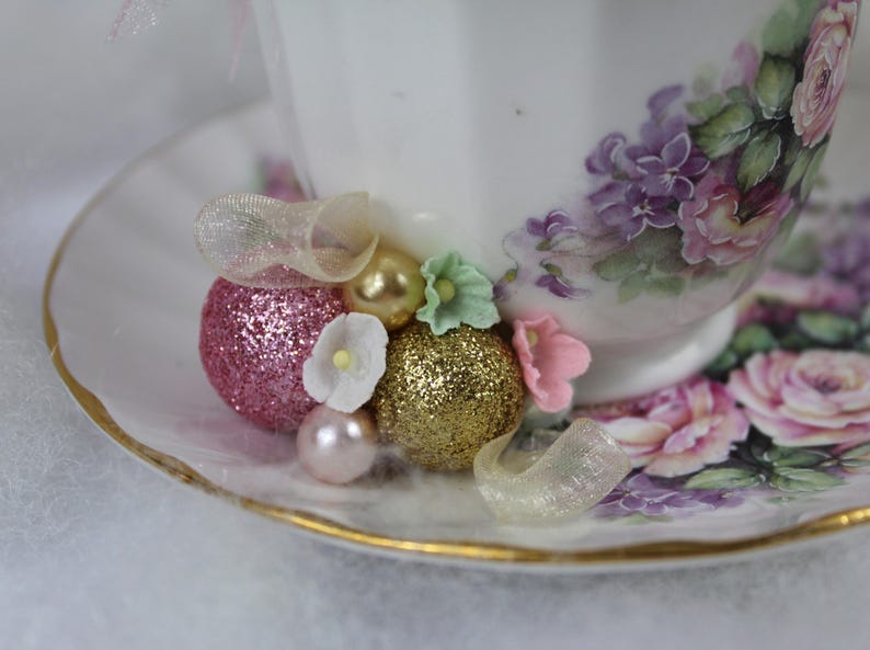 May include: A white teacup and saucer set with a floral design in pink and purple. The cup is decorated with glittery pink and gold embellishments, small white and pink flowers, and a pearl. The saucer has a gold rim.