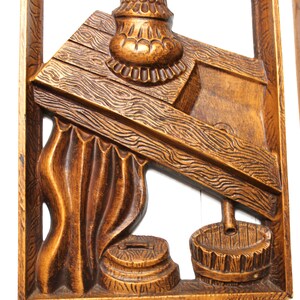 May include: A wooden wall hanging featuring a carved design of a well with a bucket and a pulley. The well is crafted from wood and has a brown finish. The bucket is made of wood and has a brown finish. The pulley is made of metal and has a silver finish.