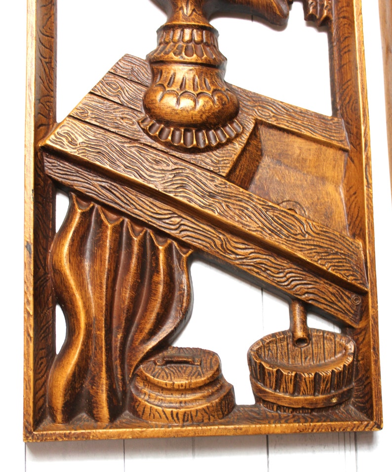 May include: A wooden wall hanging featuring a carved design of a well with a bucket and a pulley. The well is crafted from wood and has a brown finish. The bucket is made of wood and has a brown finish. The pulley is made of metal and has a silver finish.