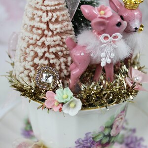 May include: A decorative teacup arrangement featuring pink deer figurines with gold crowns and floral accents. The teacup is filled with a miniature Christmas tree, gold tinsel, and a jeweled embellishment. The teacup and saucer have a floral pattern.