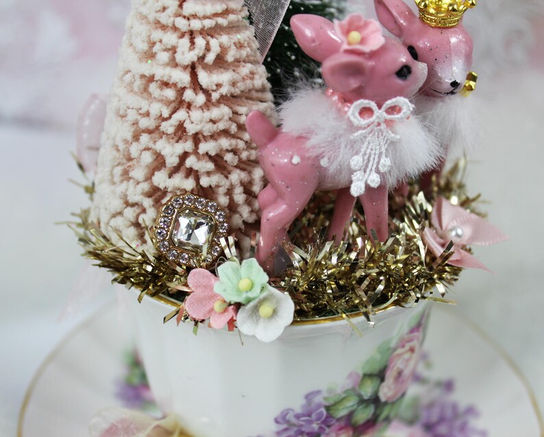 May include: A decorative teacup arrangement featuring pink deer figurines with gold crowns and floral accents. The teacup is filled with a miniature Christmas tree, gold tinsel, and a jeweled embellishment. The teacup and saucer have a floral pattern.