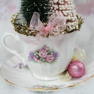 May include: A decorative teacup and saucer set with a floral design, holding miniature Christmas trees and a pink glitter ornament. The teacup is adorned with pink roses and purple flowers, and a pink bow.