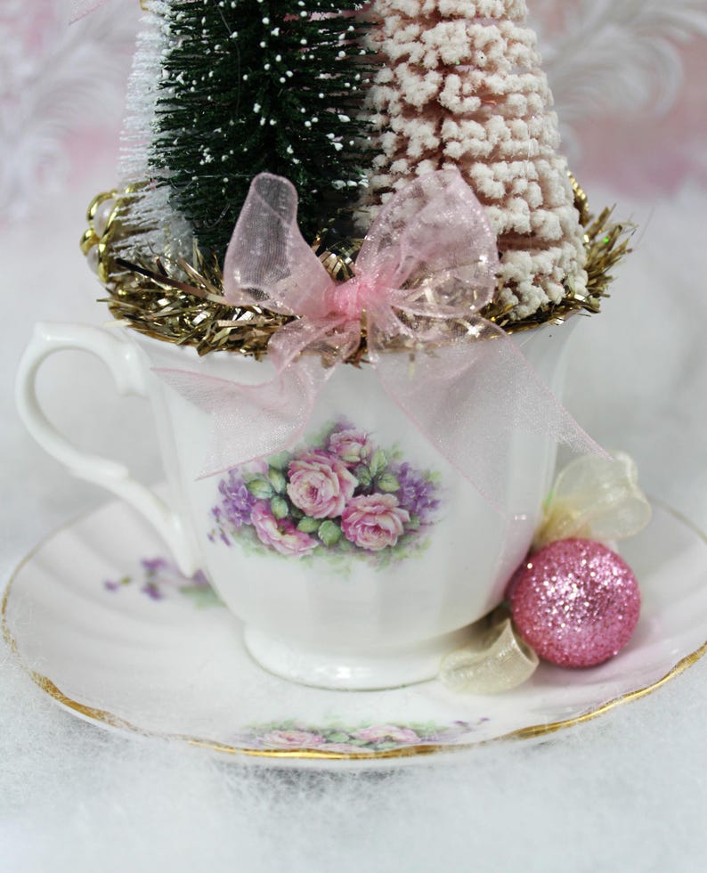 May include: A decorative teacup and saucer set with a floral design, holding miniature Christmas trees and a pink glitter ornament. The teacup is adorned with pink roses and purple flowers, and a pink bow.
