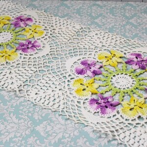 May include: A cream-colored, crocheted table runner featuring two floral designs. Each design has a central white circle surrounded by yellow and purple flowers with green leaves. The openwork lace design adds a delicate touch.
