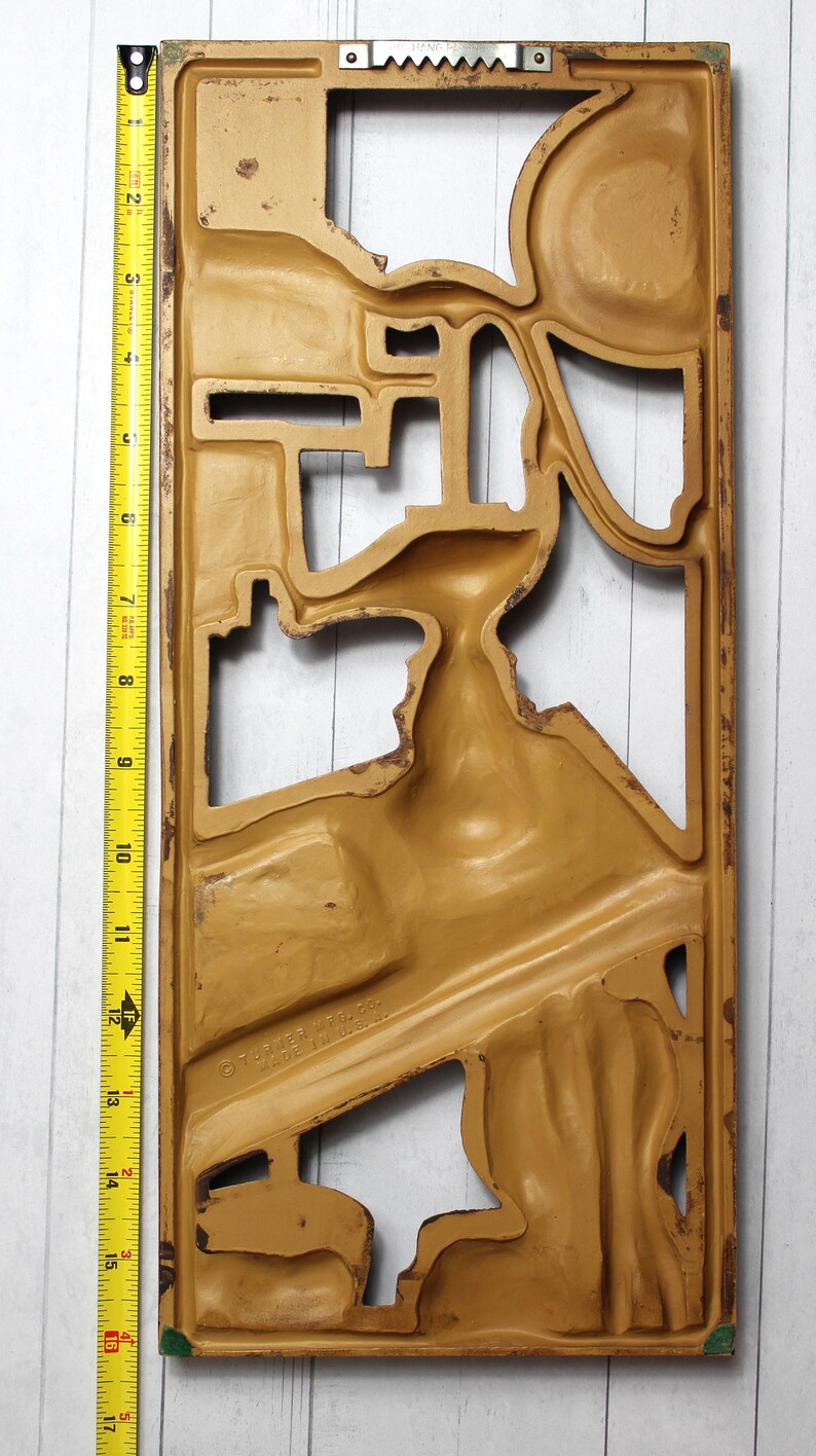 May include: A gold-coloured metal wall decor with a cutout design. The design features a series of abstract shapes and curves. The decor is approximately 17 inches tall.