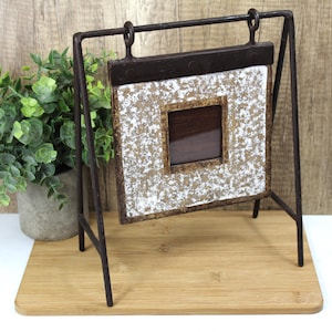 May include: A rustic, square picture frame with a distressed white and gold speckled surface, set within a dark brown frame. The frame hangs from a dark metal stand, creating a vintage aesthetic. The frame is displayed on a wooden surface.