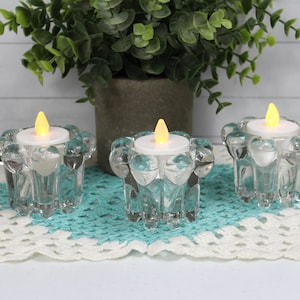 May include: Three clear glass candle holders with a textured design, each holding a white tea light candle. The candles are lit and casting a warm glow. The candle holders are sitting on a white and teal crochet doily.