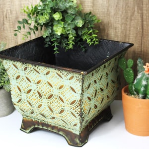 May include: Square metal planter with a distressed mint green finish and a repeating gold pattern. The planter has a dark interior and sits on four small feet. A small potted cactus and other greenery are nearby.