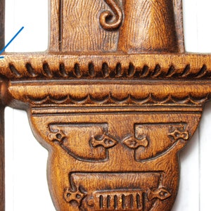 May include: A wooden wall decoration featuring a carved design of a figure holding a staff. The figure stands on a shelf with a decorative edge. The wood is a dark brown colour.
