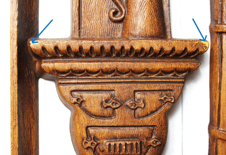 May include: A wooden wall decoration featuring a carved design of a figure holding a staff. The figure stands on a shelf with a decorative edge. The wood is a dark brown colour.
