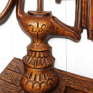 May include: A wooden wall decoration featuring a carved design of a pump and a bucket. The pump is brown and has a decorative design on the top. The bucket is also brown and has a rope handle.