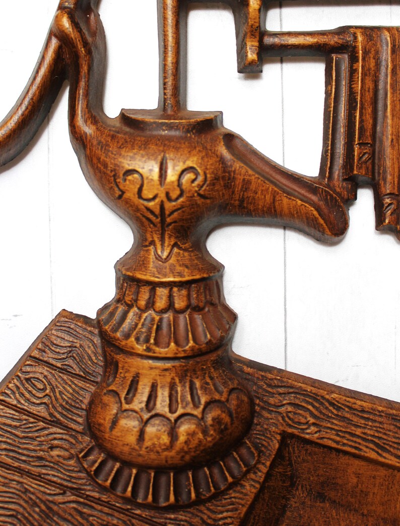 May include: A wooden wall decoration featuring a carved design of a pump and a bucket. The pump is brown and has a decorative design on the top. The bucket is also brown and has a rope handle.