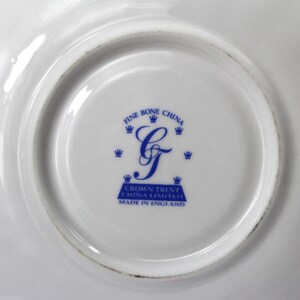 May include: Close-up of a white fine bone china plate with a blue emblem. The emblem features the words "Fine Bone China" and "Crown Trent China Limited Made in England" surrounding a stylized blue letter "G".