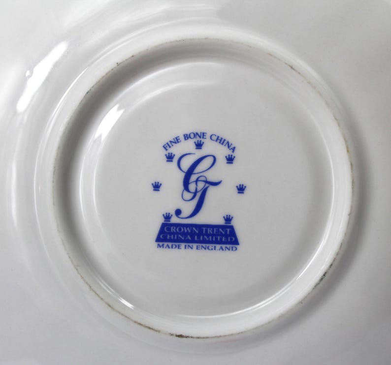May include: Close-up of a white fine bone china plate with a blue emblem. The emblem features the words "Fine Bone China" and "Crown Trent China Limited Made in England" surrounding a stylized blue letter "G".