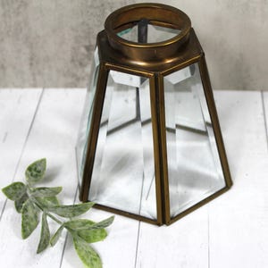 May include: A clear glass and brass lantern with a geometric design. The lantern has a hexagonal base and a cylindrical top. The glass panels are clear, and the brass frame has a warm, antique finish. A decorative green plant is in the foreground.