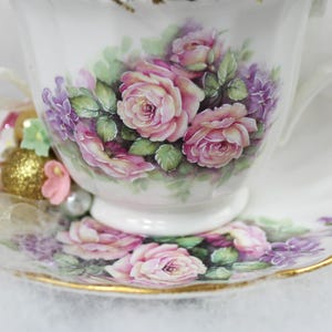 May include: White teacup and saucer set with a floral design featuring pink roses and purple flowers. The cup and saucer have gold trim. A decorative arrangement of beads and ribbons is next to the cup.
