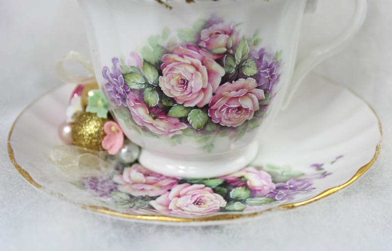 May include: White teacup and saucer set with a floral design featuring pink roses and purple flowers. The cup and saucer have gold trim. A decorative arrangement of beads and ribbons is next to the cup.