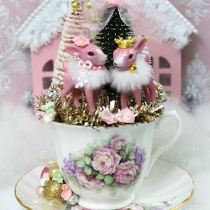 May include: A decorative teacup and saucer set with a floral design, holding a Christmas-themed arrangement. Two pink deer figurines with white fur trim and gold crowns are featured, along with miniature trees and a pink house.