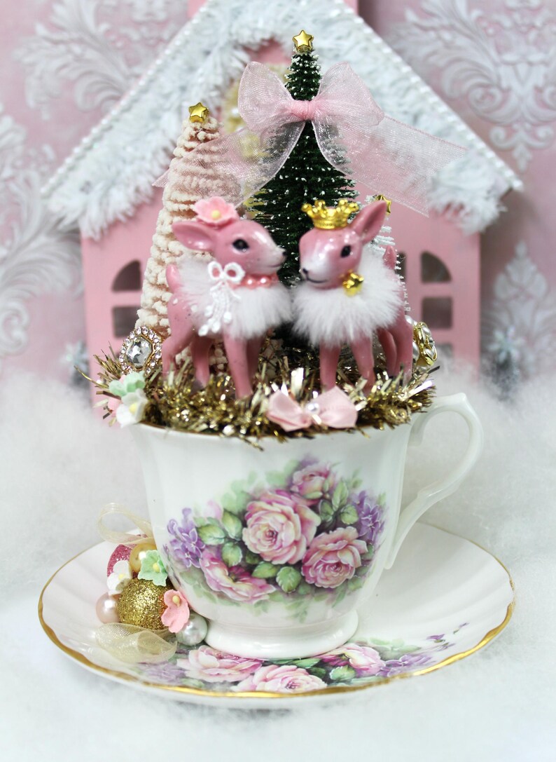 May include: A decorative teacup and saucer set with a floral design, holding a Christmas-themed arrangement. Two pink deer figurines with white fur trim and gold crowns are featured, along with miniature trees and a pink house.