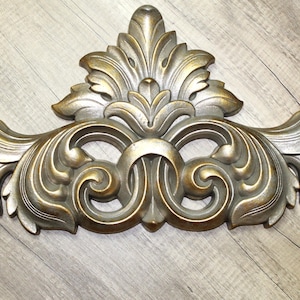 May include: Ornate, gold-toned decorative wall accent with a symmetrical design. The piece features stylized leaves and swirling patterns, creating a classic, elegant look. The wall decor is made of a durable material.
