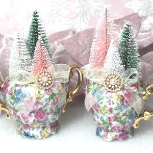 May include: Two floral porcelain sugar bowls with gold-tone handles, each decorated with a sheer ribbon and a central rhinestone brooch. Inside each bowl are miniature bottle brush trees in shades of green, white, and soft pink.