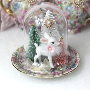 May include: A glass cloche dome sits on a floral patterned saucer, enclosing a white deer figurine with a pink flower accent. Inside are three miniature bottle brush trees in green, white, and pink, with a pearl embellishment on the white tree.