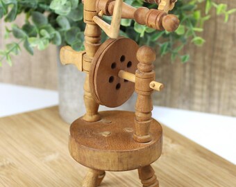 May include: A small wooden spinning wheel with a round base and three legs. The wheel is made of light brown wood and has a small spindle in the center. The wheel is decorated with a few small holes.