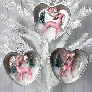 May include: Three heart-shaped Christmas decorations. Each decoration features a pink deer with gold antlers, a small Christmas tree, and a pink bow. The decorations are filled with glitter and faux snow.