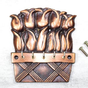May include: A bronze-colored metal key holder shaped like a basket of tulips. The key holder has five hooks and includes two screws for mounting. The tulips are detailed with curved petals.