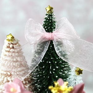 May include: Miniature Christmas trees, one green and two white, are decorated with gold star toppers and a sheer pink bow. The trees are adorned with faux snow and pink floral accents, creating a festive holiday scene.